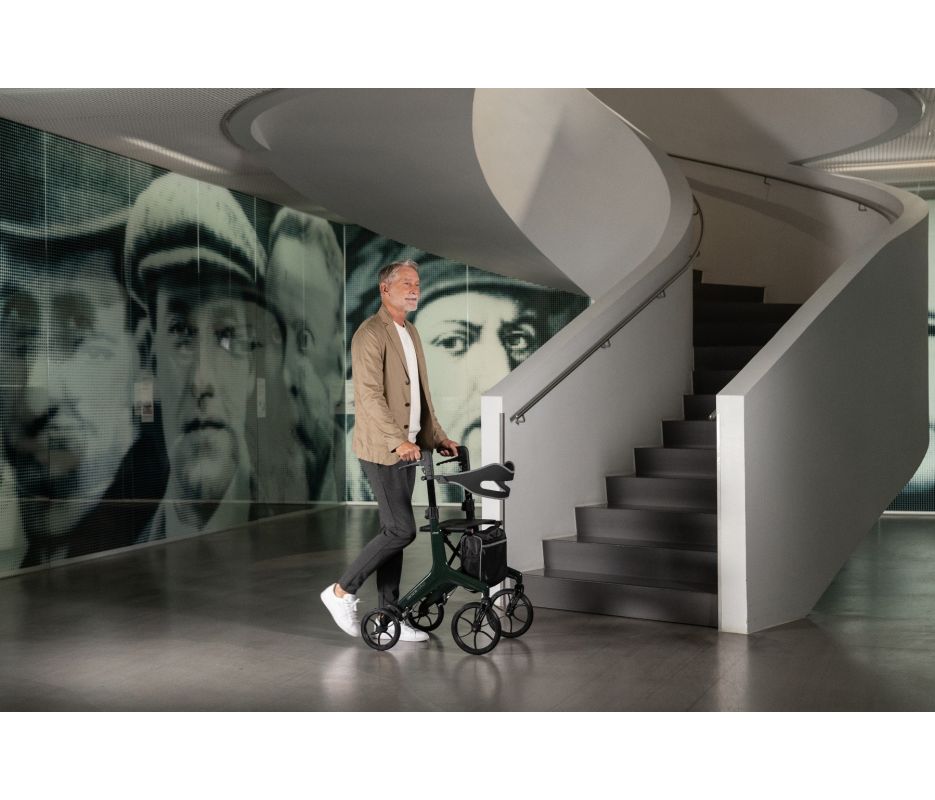 Man pushing a rollator in a modern interior setting with a staircase and wall art.