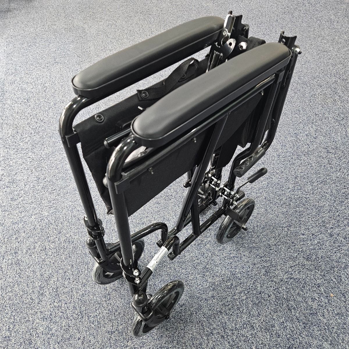 Folded black lightweight folding wheelchair with padded armrests and small rear wheels, positioned on a blue carpeted floor.