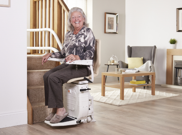 Smiling woman using a stairlift to travel safely upstairs at home, demonstrating how a stairlift supports independent living, comfort and everyday mobility.
