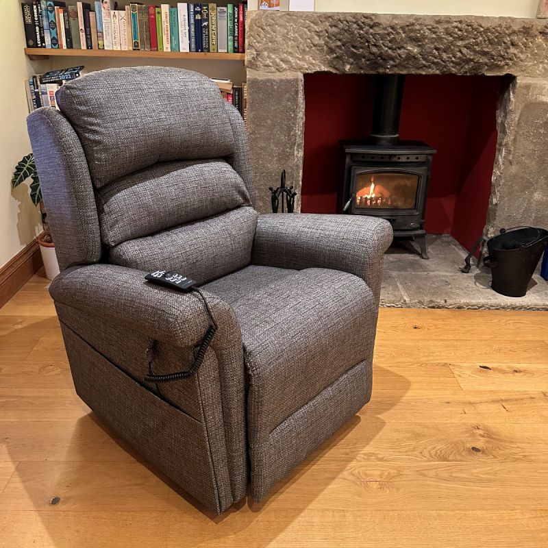 Grey fabric recliner chair with remote control, positioned on a wooden floor in front of a lit wood-burning stove and stone fireplace.