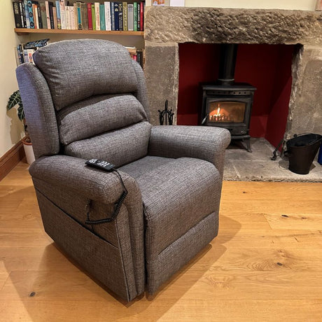 Grey fabric recliner chair with remote control, positioned on a wooden floor in front of a lit wood-burning stove and stone fireplace.