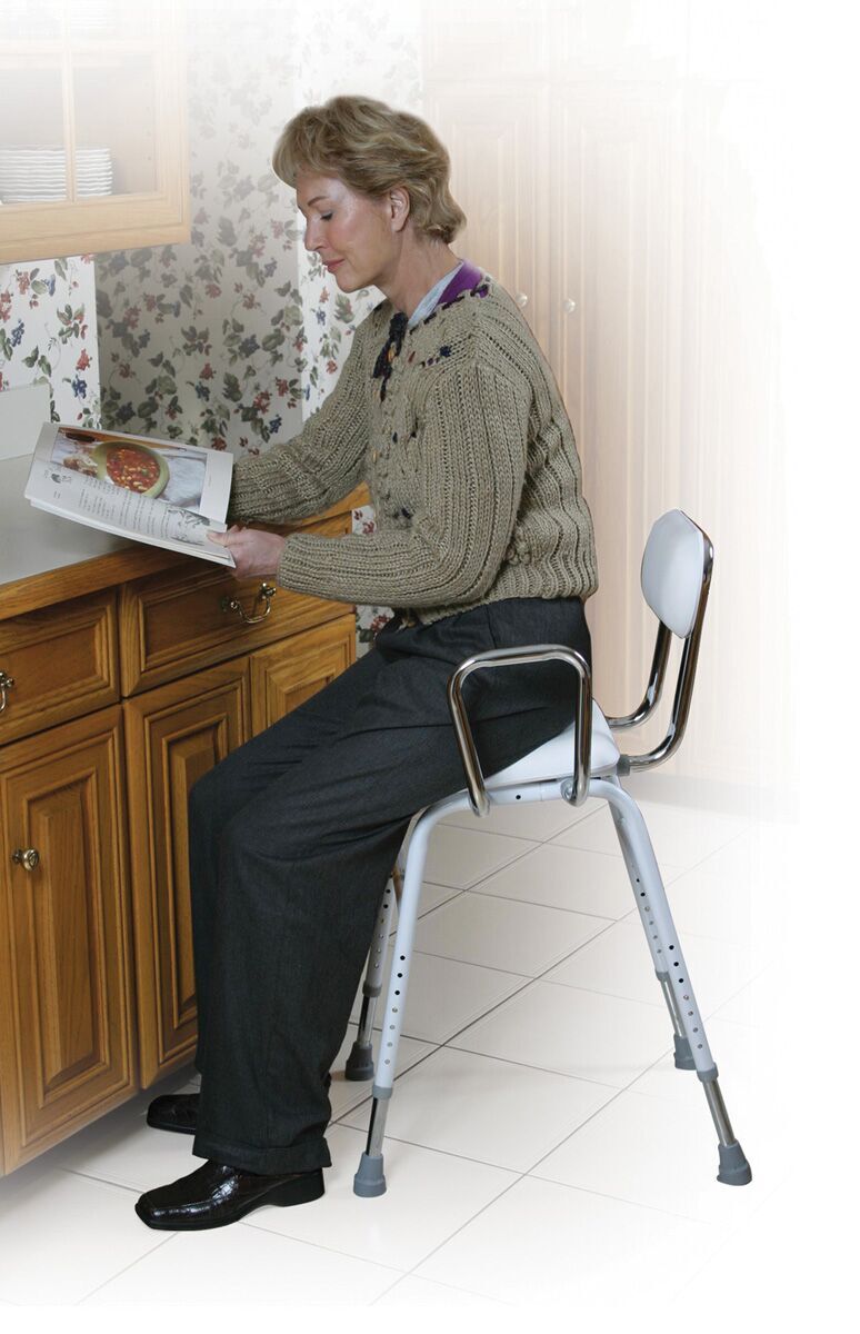Older woman sitting on a perching stool while reading a magazine in a kitchen with wooden cabinets and floral wallpaper.