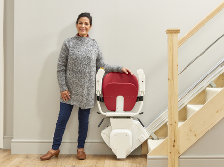 Woman standing beside a stairlift for home installed on a wooden staircase, showcasing a compact and accessible mobility solution.