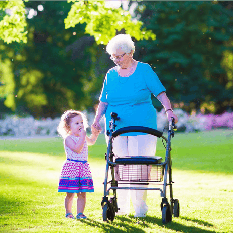 Elderly woman using a walking frame while holding hands with a young girl in a sunny park.