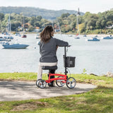 Person using a red knee walker by a lakeside with boats and trees in the background