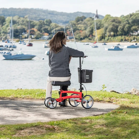 Person using a red knee walker by a lakeside with boats and trees in the background