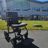 Black electric wheelchair in front of a building with 'Fenetic Wellbeing' branding.