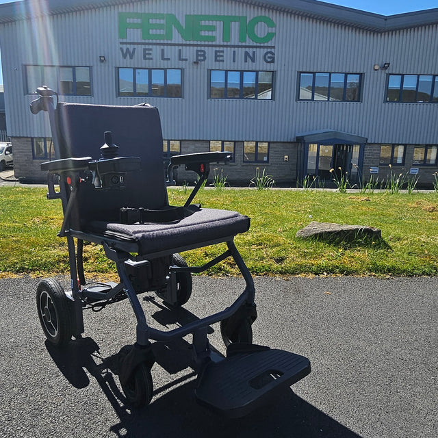 Black electric wheelchair in front of a building with 'Fenetic Wellbeing' branding.