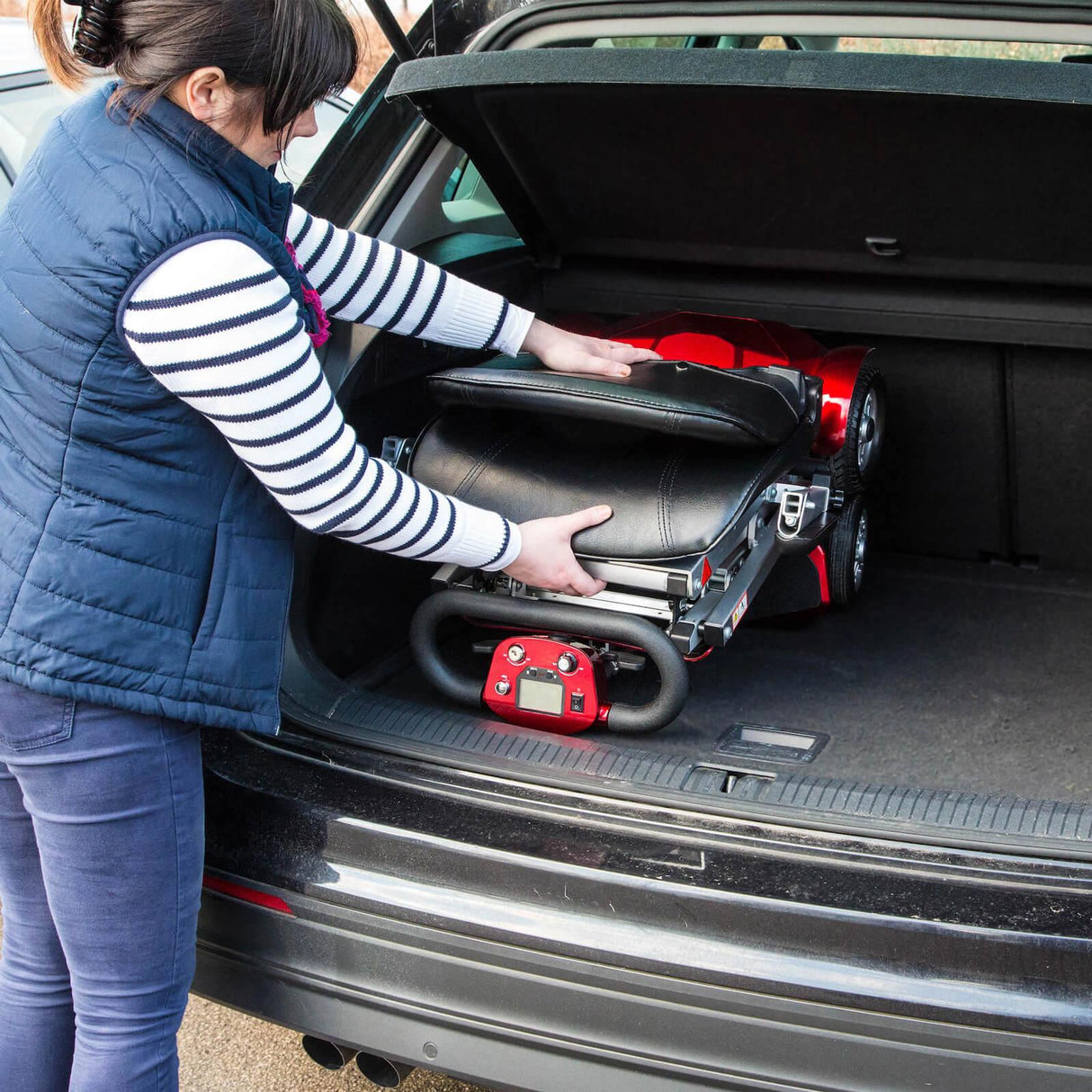 Person loading a red and black mobility scooter into a car boot. Demonstrating the compact folding size.