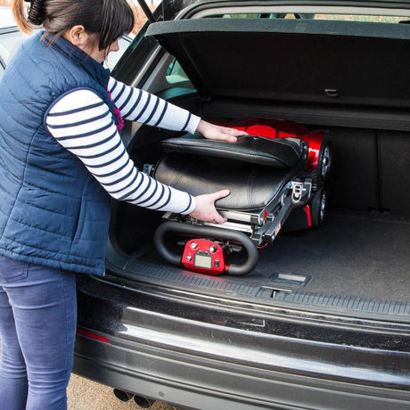 Person loading a red and black mobility scooter into a car boot. Demonstrating the compact folding size.