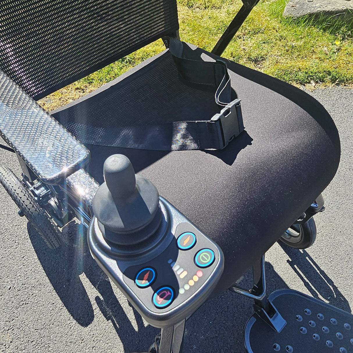 Close-up of a control panel on a powerchair with grass and pavement in the background.