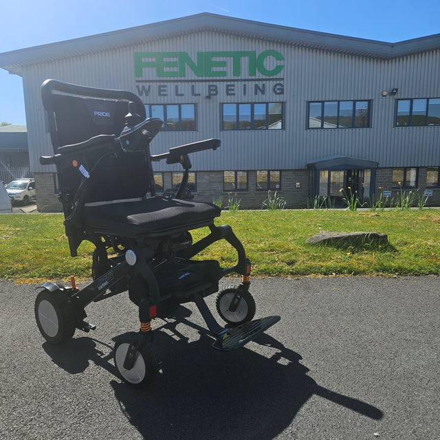 Black electric wheelchair in front of a building with 'Fenetic Wellbeing' branding.